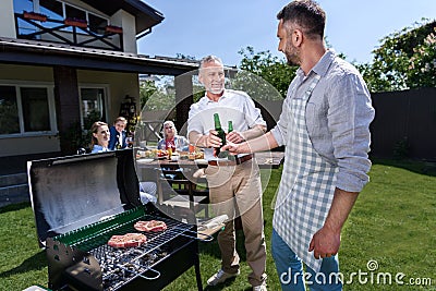 Senior father and adult son drinking beer while grilling meat outdoors Stock Photo