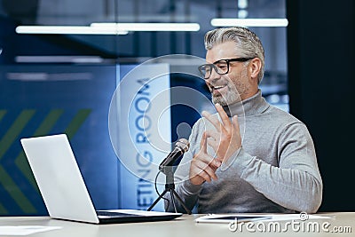 Senior man sitting an office with a microphone in front of a laptop. Talks on a video call Stock Photo