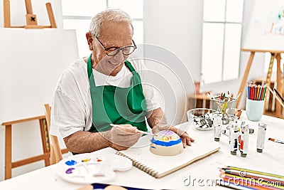 Senior grey-haired artist man smiling happy painting pottery sitting on the table at art studio Stock Photo