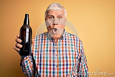 Senior handsome hoary man drinking bottle of beer standing over isolated yellow background scared in shock with a surprise face, Stock Photo