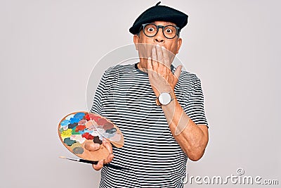 Senior hoary artist man wearing french beret drawing using paintbrush and palette with colors cover mouth with hand shocked with Stock Photo
