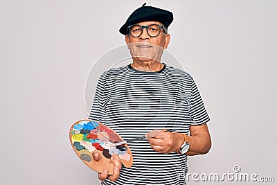 Senior hoary artist man wearing french beret drawing using paintbrush and palette with colors with a happy face standing and Stock Photo