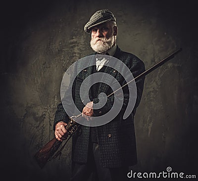 Senior hunter with a shotgun in a traditional shooting clothing, posing on a dark background. Stock Photo