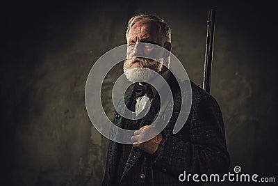 Senior hunter with a shotgun in a traditional shooting clothing, posing on a dark background. Stock Photo