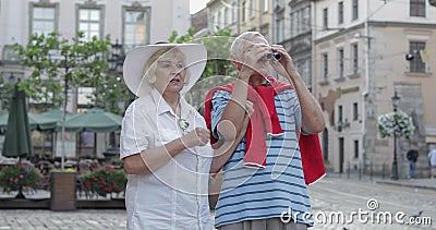 Senior male and female tourists walking in town center and looking in binoculars. Senior two male and female tourists walking in town center while traveling in stock footage Senior male and female tourists walking in town center and looking in binoculars. Senior two male and female tourists walking in town center while traveling in stock footage