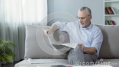 Senior male sitting on sofa and reading newspaper, press and news, rest time Stock Photo