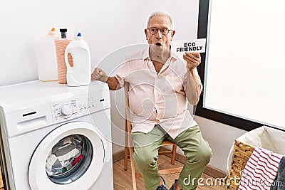 Senior man doing laundry holding eco friendly paper scared and amazed with open mouth for surprise, disbelief face Stock Photo