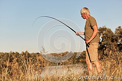 Senior man fishing, holding fishing pole in hands, wearing green t shirt and short, looking at bobber and pulling out fish, Stock Photo