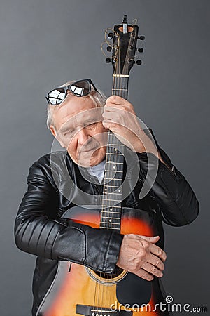 Senior man in leather jacket and sunglasses up on head standing isolated on gray hugging guitar close closed eyes Stock Photo