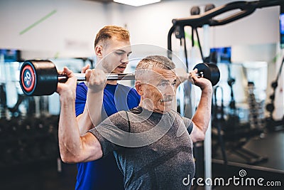 Senior man lifting weights with help of gym assistant. Stock Photo