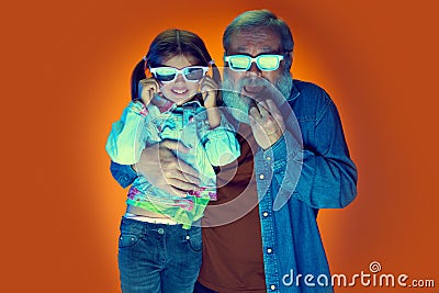 Senior man and little girl, grandfather and granddaughter watching TV over gradient orange background in neon light Stock Photo