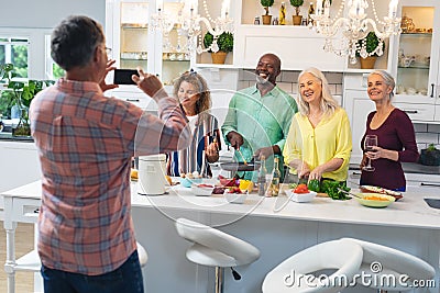 Senior man photographing smiling multiracial male and female friends preparing food at home Stock Photo