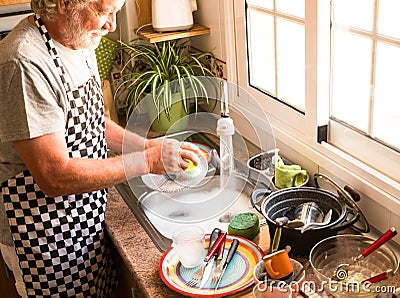 A senior man white hair and beard wearing apron and gloves washing dishes. Corner of kitchen. Grandfather at work. Bright light Stock Photo