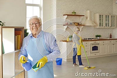 Senior man wipes the shelves holding a washcloth and detergent while his wife washes the floor. Stock Photo