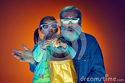 Senior man and little girl, grandfather and granddaughter watching TV over gradient orange background in neon light Stock Photo