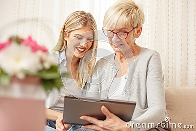 Senior mother and daughter smiling and looking at the tablet. Flower bouquet in foreground. Stock Photo
