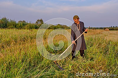An senior Muslim man in a skullcap and traditional clothes mows hand-scythe grass in a hayfield Stock Photo