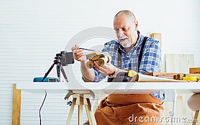 Senior old Caucasian man wearing check shirt, apron, making DIY wooden furniture, using mobile phone, streaming live video clip, Stock Photo