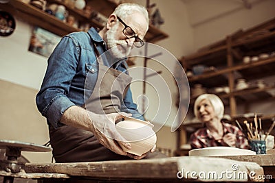 Senior potter in apron and eyeglasses examining ceramic bowl with woman working Stock Photo