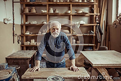 Senior potter standing and leaning on table against shelves with pottery goods at workshop Stock Photo