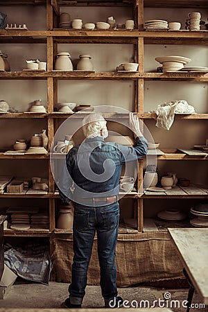 Senior potter standing near shelves with pottery goods and searching something at workshop Stock Photo