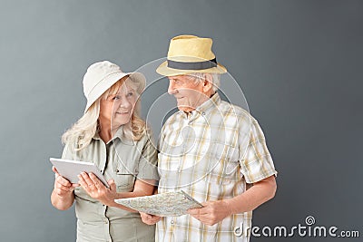 Senior tourists in beach hats studio standing isolated on gray holding map using digital tablet looking at each other Stock Photo