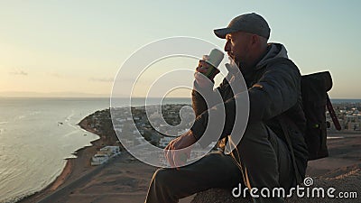 Senior traveler open thermocup and drinking hot drink while sitting on a hill Stock Photo