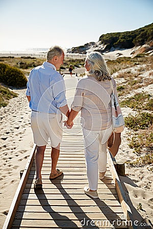 Senior white couple walking along wooden promenade on a beach holding hands, full length, back view Stock Photo