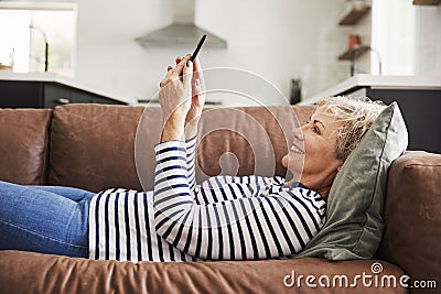 Senior white woman lying on couch at home using smartphone Stock Photo