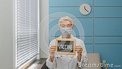 Senior woman doctor with disposable mask holds blackboard with word Vaccine Stock Photo