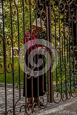 Senior woman in dress and hat locks wrought iron gate at property entrance with chain and padlock - lush vegetation and Editorial Stock Photo