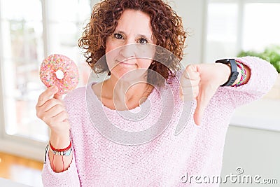 Senior woman eating pink sugar donut with angry face, negative sign showing dislike with thumbs down, rejection concept Stock Photo