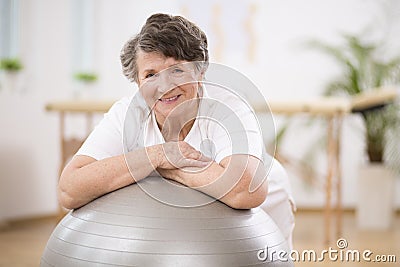 Senior woman leaning on grey gymnastic ball at physiotherapy center Stock Photo