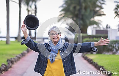 Senior woman. Smiling elderly female grey haired enjoying winter sea with raised arms and black hat in the hand. Active carefree Stock Photo