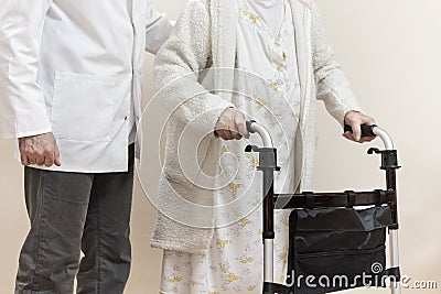 Senior woman in a white bathrobe and nightgown goes with a rehabilitation walker. The nurse supports and protects the old wom Stock Photo