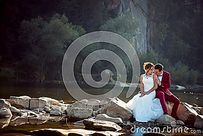 Sensitive portrait of the groom in red suit kissing the adorable bride in the shoulder while sitting on the stone near Stock Photo