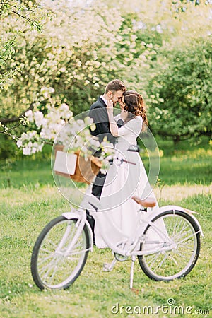 Sensual young newlywed couple holding each other in park. Bicycle with wedding decoration on foreground Stock Photo