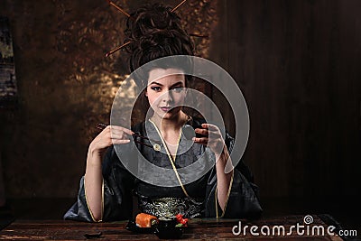 Sensual young woman in a geisha asian costume with fashion makeup and hair style drinks tea and eats sushi Stock Photo