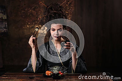 Sensual young woman in a geisha asian costume with fashion makeup and hair style drinks tea and eats sushi Stock Photo