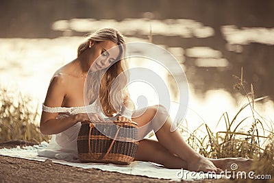 Sensual young woman on the picnic sitting by the lake at sunset and looking in picnic basket Stock Photo