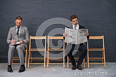 Separating the competition. Shot of a row of chairs with two businessmen seated slightly apart. Stock Photo