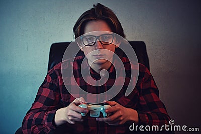 Serious adolescent boy playing video games late night seated in armchair inside a dark room background. Intent teen guy holding a Stock Photo