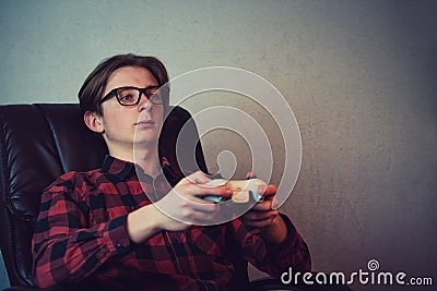 Serious adolescent boy playing video games late night seated relaxed in his armchair over grey wall background. Intent teen guy Stock Photo