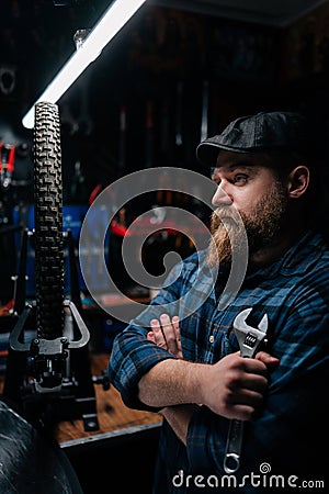 Serious bearded cycling repairman in cap holding wrench standing with crossed arms by bicycle in repair workshop with Stock Photo