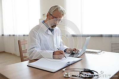 Serious busy mature doctor filling documents, sitting at work desk Stock Photo