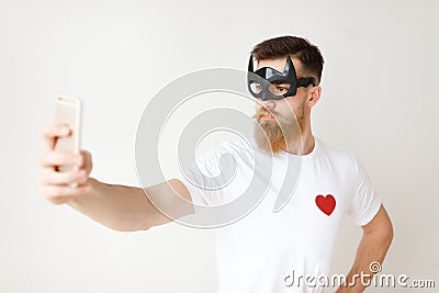 Serious confident bearded male wears batman mask, poses in front of smart phone camera, makes selfie, feels to be hero Stock Photo