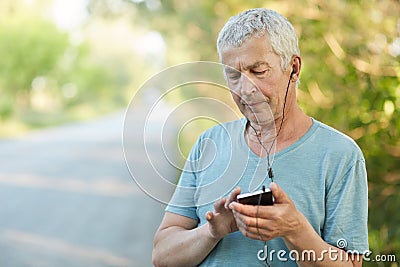 Serious elderly wrinkled male uses smart phone and earphones for listening music, focused into screen, has stroll in countryside, Stock Photo