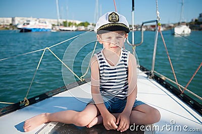 Serious kid in captain hat sitting on luxury yacht board in sea port in summer Stock Photo