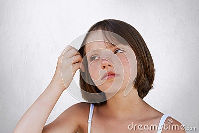 Serious little freckled child, keeping her hand on hair, havin thoughtful expression, looking aside. Beautiful girl posing against Stock Photo