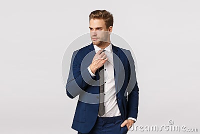 Serious-looking sassy businessman in blue classis suit, adjusting tie and looking away, holding hand in pocket, getting Stock Photo